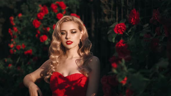 Fabulous Girl with Red Lips in Red Dress on Awesome Summer Roses Background. Fantasy Woman Portrait