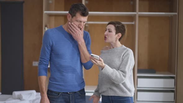 Furious Jealous Beautiful Wife Showing Smartphone Yelling at Sad Husband Standing in Bedroom at Home