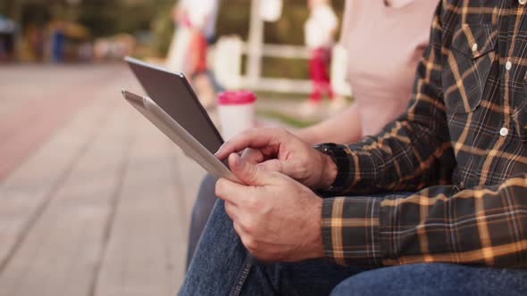 Man is Sitting on Park Bench with Tablet in His Hands and Typing Something on It Closeup Side View