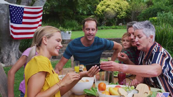 Smiling caucasian family having celebration meal in garden looking at smartphone together
