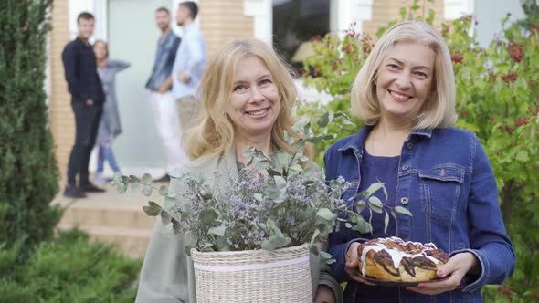 Two Caucasian Mature Women Smiling and Holding a Cake and a Bucket of Flowers