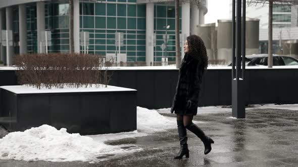 A Darkhaired Curlyhaired Woman in a Fur Coat and Highheeled Boots Is Walking Around the Winter City