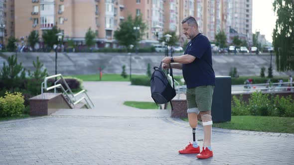 Side View Portrait Caucasian Man with Artificial Legs Hanging Backpack on Shoulder Walking in Urban