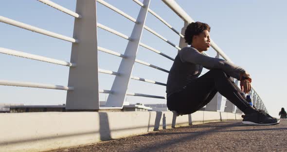 Fit african american man exercising outdoors in city, resting sitting on footbridge