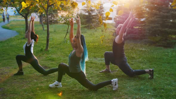 Two Young Women Doing Yoga Asanas with Trainer in the Park in the Rays of the Sun