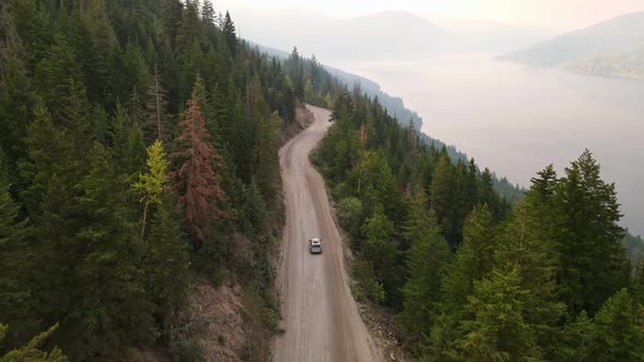 Silver van driving along a dusty forest service road during wildfire season with Adams Lake in the b