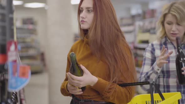 Gorgeous Redhead Woman Choosing Drink on Supermarket Shelves As Her Blond Friend Coming with Bottle