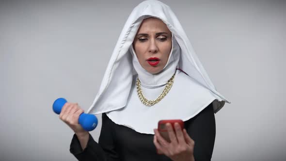 Absorbed Caucasian Woman in Nun Costume Lifting Dumbbell Watching Online Lesson in Smartphone
