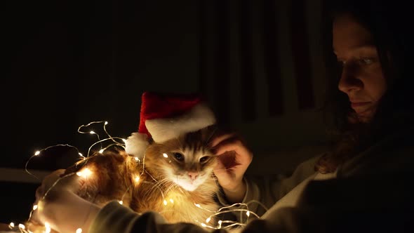 Woman Holds Cat in Christmas Hat and Wrapped with Garland