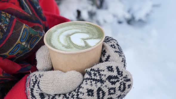 Hands in Knitted Mittens holding Steaming Cup of Hot Coffee or Tea on Snowy Winter Morning