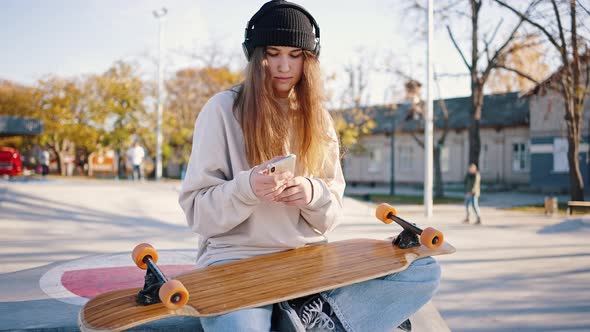 Outside Portrait of a Schoolgirl with a Skateboard and a Smartphone in a Skatepark Listening to