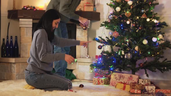 Man and Woman Decorating Christmas Tree in Modern Room. Girl Gives Toys, Man Hanging Them on