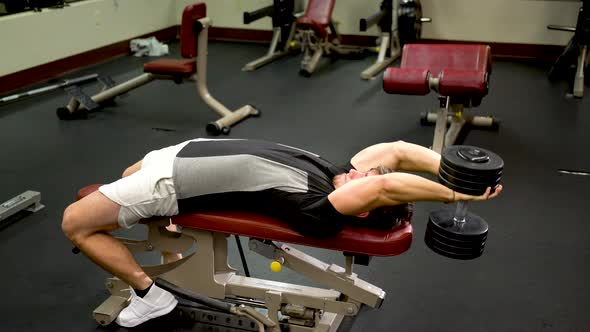 Side shot of teen bodybuilder lifting a heavy dumbbell over his head while lying on a bench in a gym