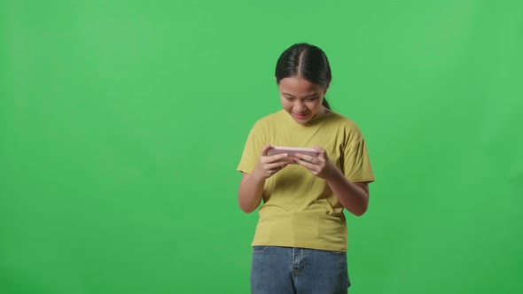 Excited Asian Kid Girl Playing Video Game With Mobile Phone In Green Screen Studio