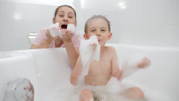 Happy Laughing Boy with Mother Playing in Bath and Blowing Soap Foam and Bubbles in Camera