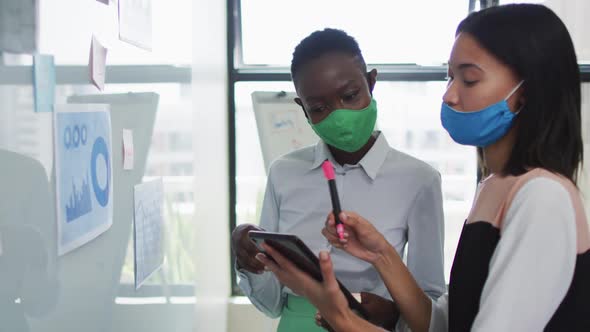 Diverse female office colleagues wearing face masks discussing over digital tablet at modern office