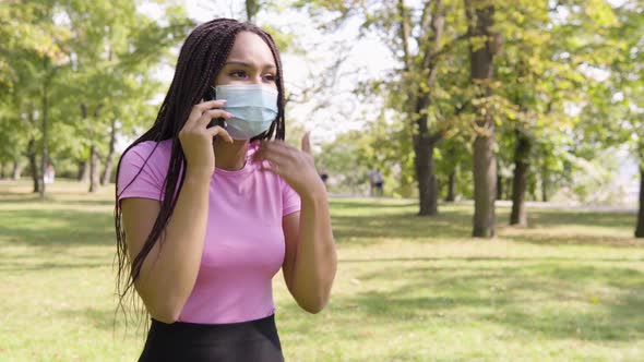 A Young Black Woman in a Face Mask Argues with Someone on a Smartphone in a Park on a Sunny Day