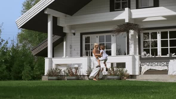 Happy Young Mother Having Fun with Her Son Taking Him in His Arms Near the House