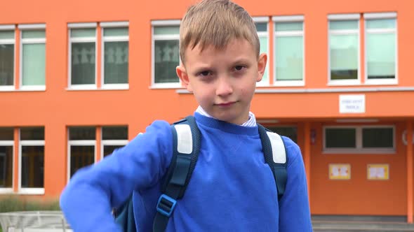 A Young Boy Frowns and Shows a Thumb Down To the Camera - an Elementary School in the Background