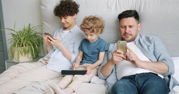 Man and Woman Using Smartphones While Kid Playing Game on Tablet in Bed at Home