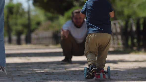 Little Boy Skating on the Scooter to His Father in the Park