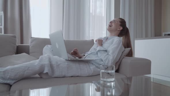 Young Woman Wearing Pajamas Lying on Sofa Watching on Computer - Happy Girl Having Fun Using Laptop