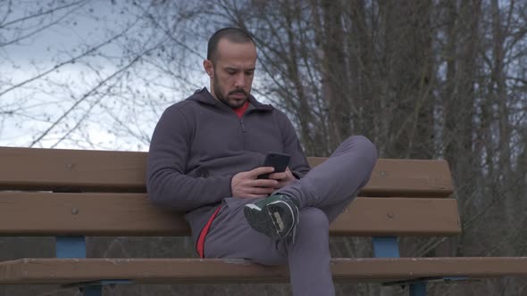 Closer shot of young bearded man with short hair sitting on a park bench on a cloudy fall day and pl