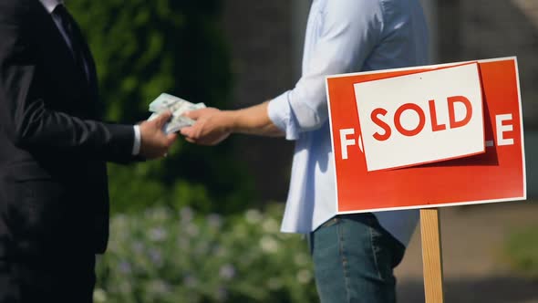 Estate Agent and Man Exchanging Money and House Keys Against Sold Signboard