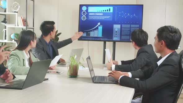 Young Asian Businessman Presentation To Business Colleagues, Pointing At Charts On TV