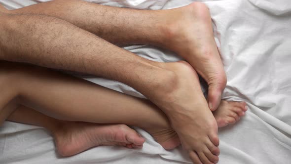 Top view. Female and male legs in bed on a white sheet caress each other. Romance at home