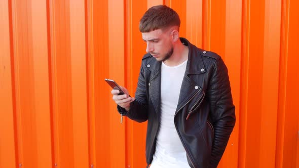 Young Man with a Beard in a Black Jacket Looks Into the Phone on an Orange Background, Slo-mo