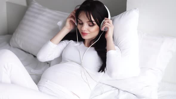 Pregnant Woman Listening Music In Headphones And Sitting On Bed