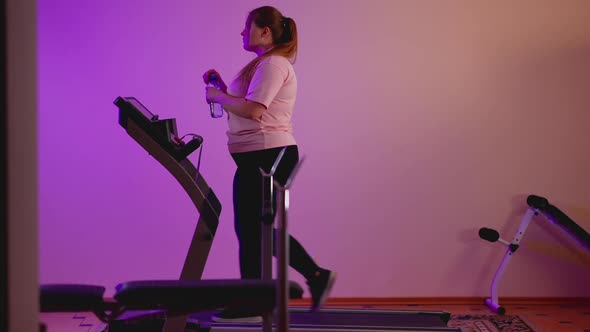 Side View of Plump Caucasian Woman Walking on Treadmill Drinking Refreshing Water