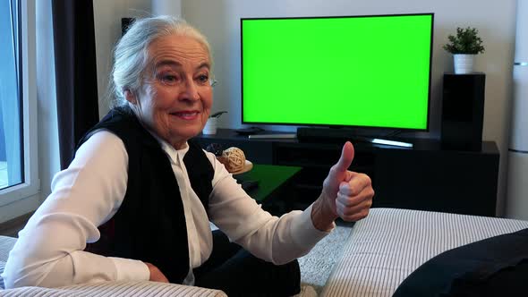 Old Caucasian Woman Watches Television in Living Room and Then Shows Thumb Up