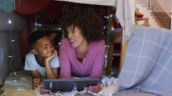 Happy african american mother and son lying in house made of blankets, using tablet