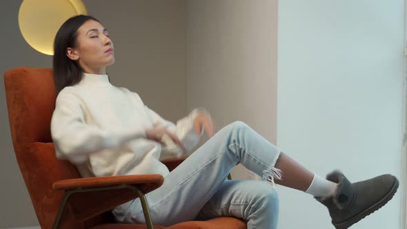 Asian Woman in a White Sweater Sitting Comfortably in an Armchair Throws Her Hands Behind Her Head