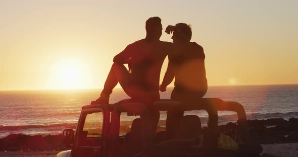 Happy caucasian gay male couple sitting on car roof embracing at sunset on the beach