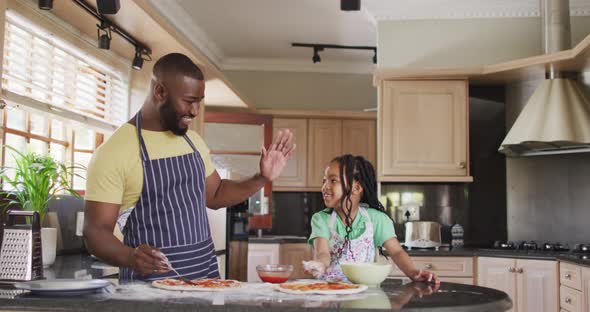 Happy african american father and daughter baking, preparing pizza in kitchen and clapping hands