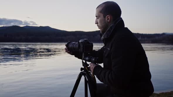 Caucasian man adds filter to camera lens attached to tripod by calm lake waters with dark mountain r