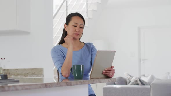 Focused asian woman sitting at table, drinking coffee and using tablet in kitchen