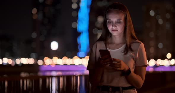 A Young Girl on the Background of the Night City Looks Into the Smartphone Screen