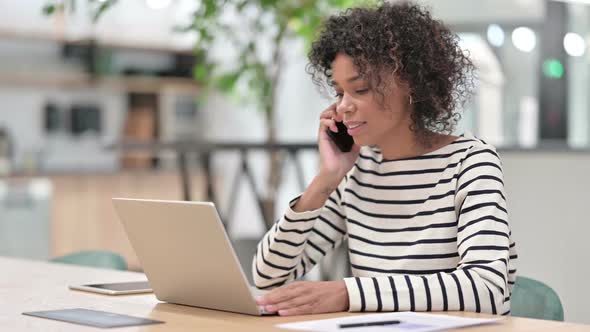 African Woman with Laptop Talking on Smartphone in Office