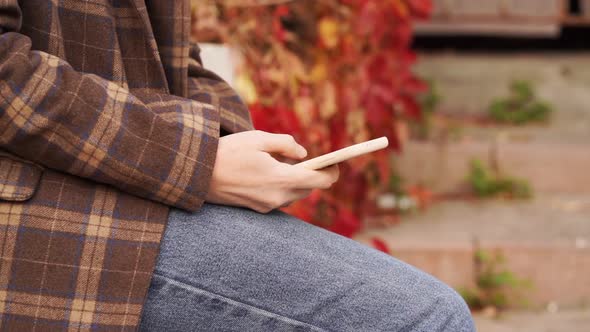 a Woman Holds a Smartphone in Hands and Views Social Media Feed in Autumn Park