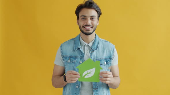 Smiling Middle Eastern Man Holding Green Energy Symbol House with Leaf on Yellow Background