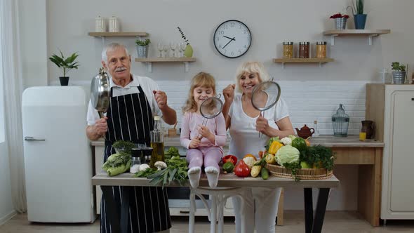 Happy Vegan Senior Couple Dancing with Granddaughter Child While Cooking Vegetables in Kitchen