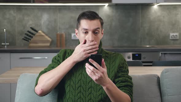 Shocked Man Reading News on His Smartphone While Sitting on Sofa at Home