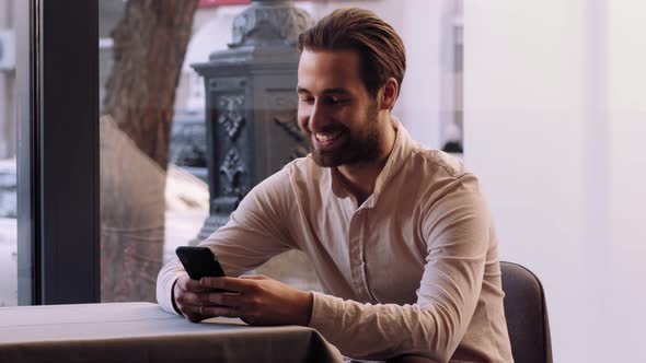 Cheerful Man Browsing on Cellphone Sitting at Table in Cafe Waiting for Date