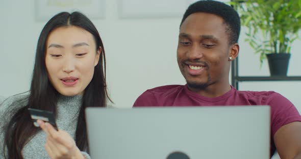 Portrait of Mixed Ethnicity Couple Sitting on Sofa in Living Room Browsing on Laptop Buying on