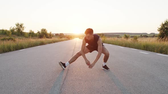 Handsome Athlete with Sportswear Warming Up Legs on Country Road with Sunset