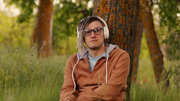 Young Relaxed Man in Glasses Headphones Sitting Near Tree in Green Grass Listening to Music on